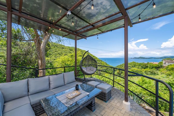 Outdoor patio with gray sofa, hanging chair, table, string lights, ocean and greenery view under a clear sky.