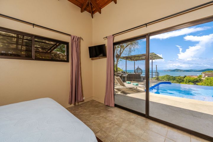 Room with bed, TV, sliding glass doors opening to pool and ocean view.