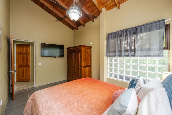 Bedroom with orange bedding, wooden wardrobe, wall-mounted TV, and large windows with sheer curtains.
