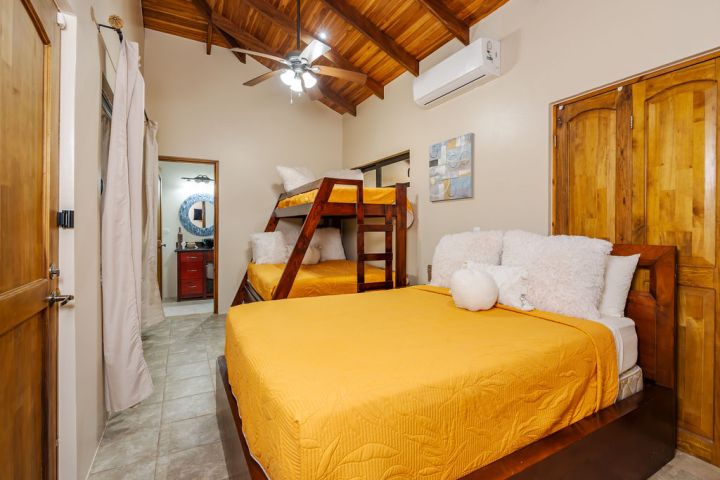 Cozy bedroom with bunk bed, main bed, and wooden ceiling, features an air conditioner and open bathroom door.