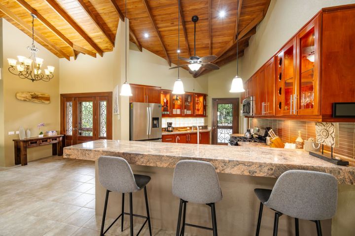 Spacious kitchen with wooden cabinets, a marble island, and pendant lighting. Three gray chairs are at the counter.