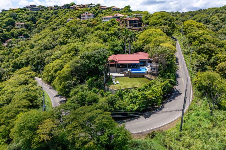 A hilltop house with a red roof and pool, surrounded by lush greenery and a winding road.