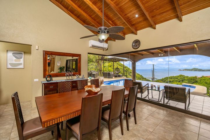 Dining room with wooden table and ocean view through large glass doors leading to a terrace with pool.
