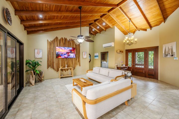 Spacious living room with wooden ceiling, light sofa, TV, chandelier, and large doors.