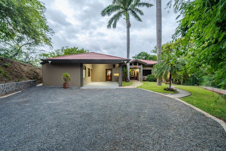 Single-story house with red roof, surrounded by palm trees and a gravel driveway.