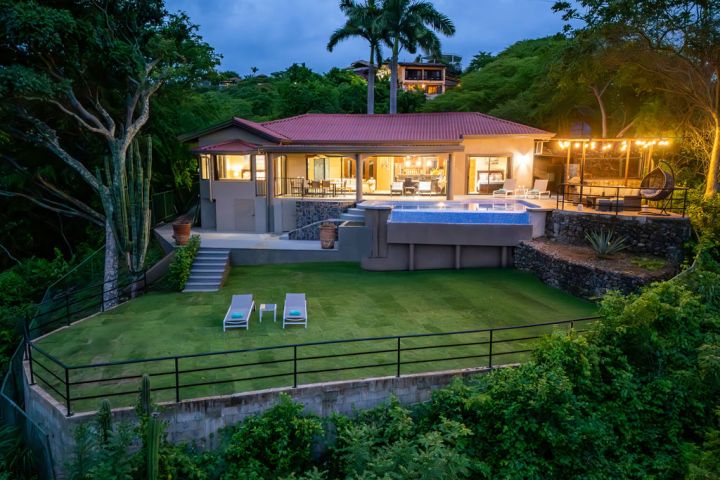 Modern house with a pool, palm trees, and outdoor lights on a hillside at dusk.