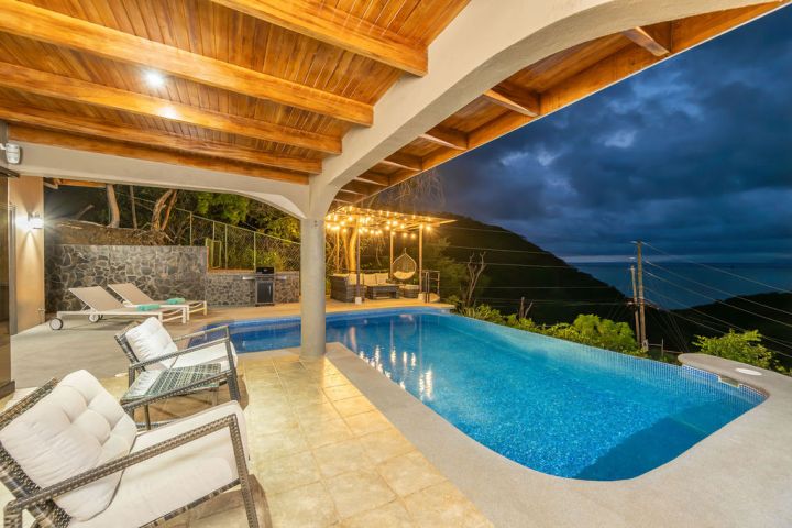 Covered patio with seating, pool, ocean view at dusk.