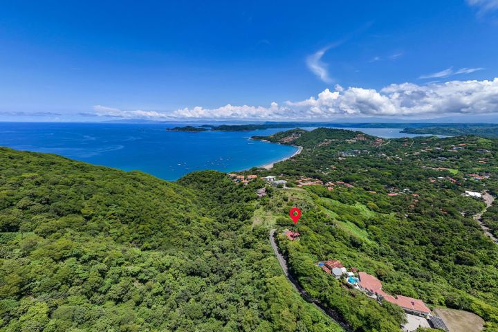 Aerial view of a lush coastline with bay, hills, and clouds under a blue sky.