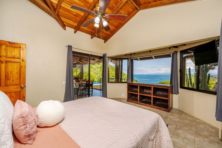 Bedroom with wooden ceiling, bed, TV, ocean view through large windows, and door to balcony.