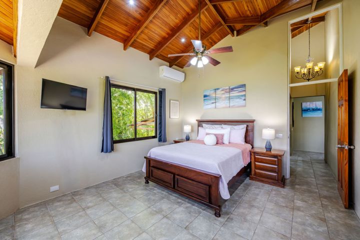 Spacious bedroom with wooden ceiling, bed, TV, and windows, featuring ceiling fan and chandelier.