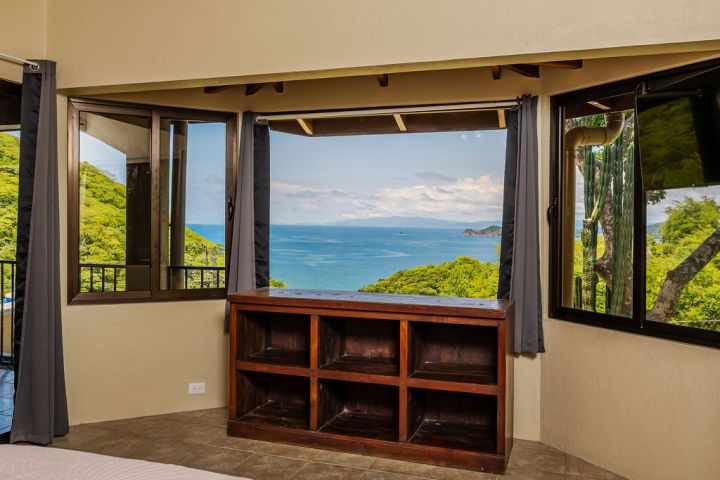Bedroom with ocean view, wooden ceiling, fan, and empty shelf unit under large window.