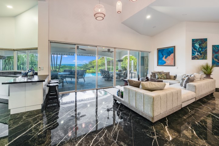 Spacious living room with marble floors, large sofa, art on walls, opens to a patio with pool and scenic view.