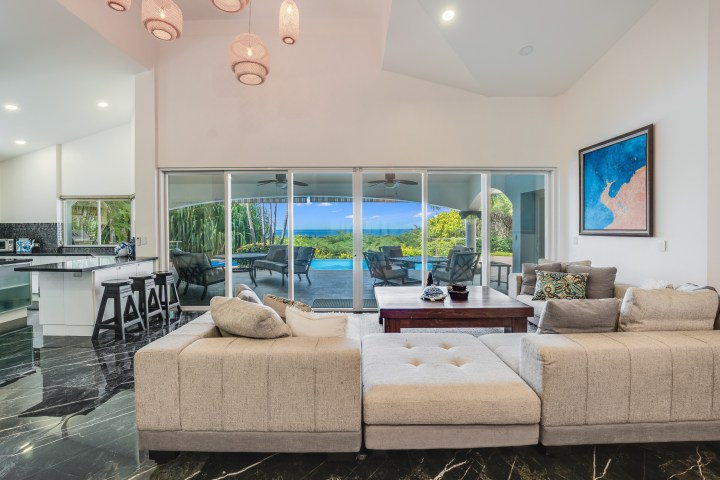 Spacious living room with beige sofas, sliding glass doors, ocean view, and adjacent kitchen. Marble flooring.