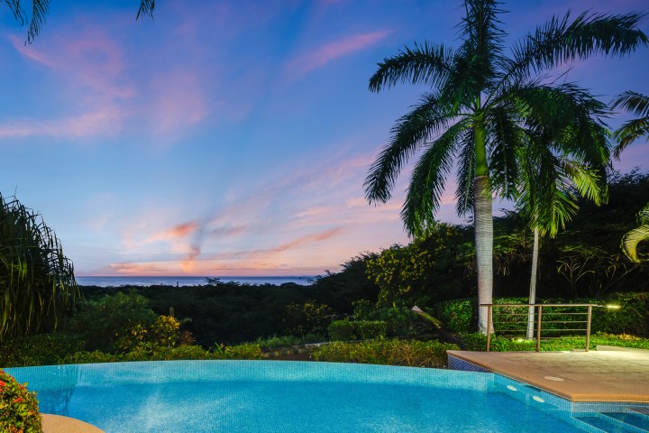 Tropical sunset over an infinity pool with palm trees and ocean in the background.