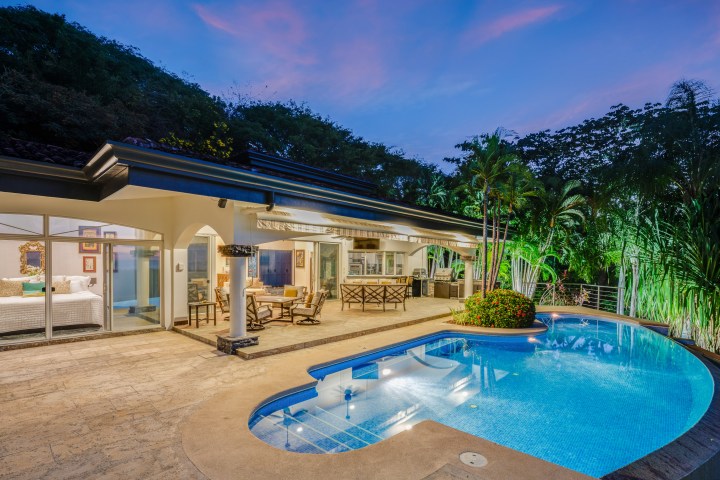 Modern house with pool, patio, and outdoor dining area at dusk, surrounded by lush greenery.