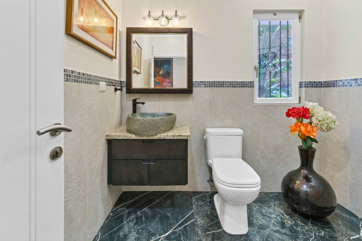 Modern bathroom with vessel sink, framed mirror, toilet, and vase of flowers on black tile floor.