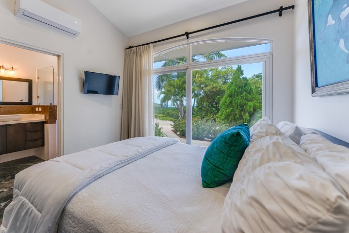Bedroom with large window, bed, TV, and view of bathroom and garden.