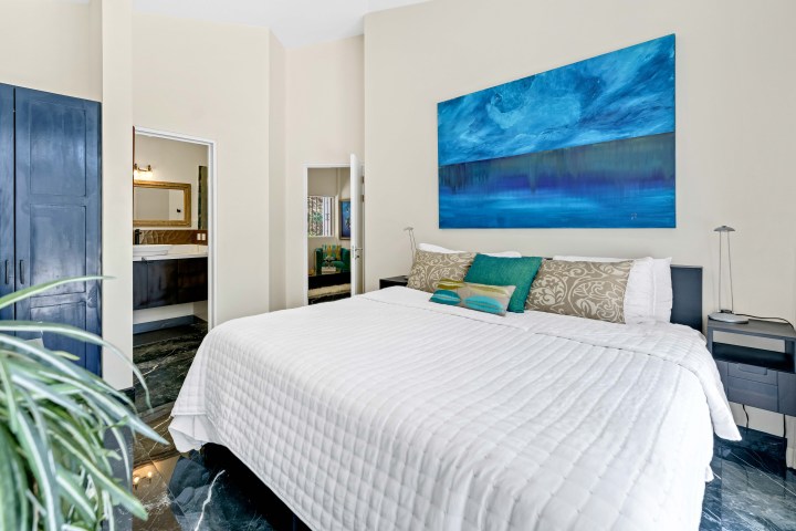 Bright bedroom with a large bed, blue artwork, plants, and a view into a modern bathroom.