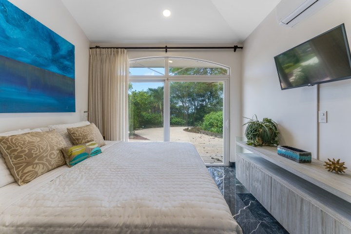 Modern bedroom with large bed, abstract art, TV, and glass doors opening to a patio with trees.