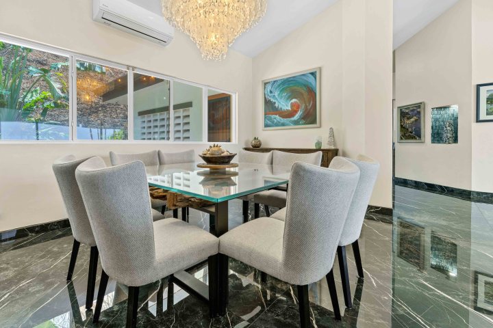 Dining room with a glass table, eight chairs, chandelier, and ocean wave painting on the wall.