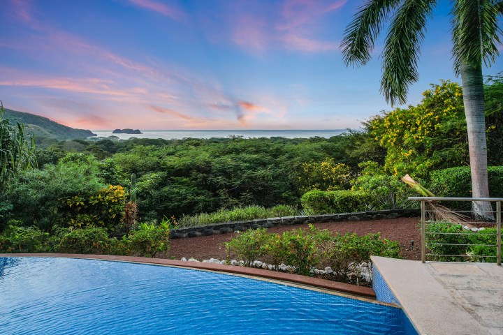 Infinity pool overlooking lush greenery and ocean at sunset, with palm trees and distant islands visible.