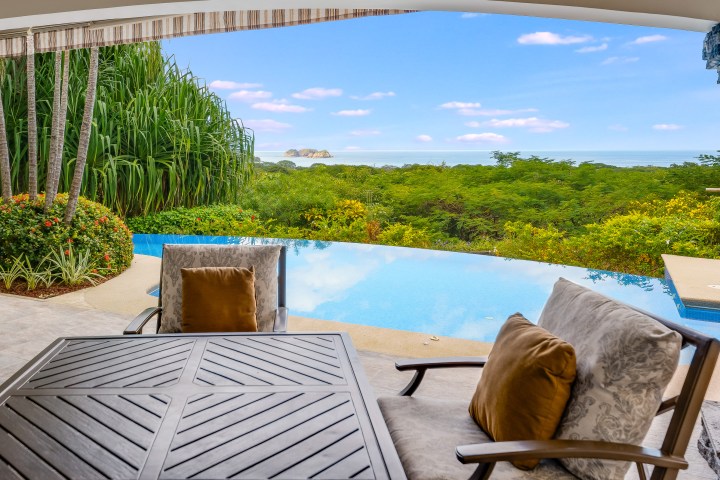 Patio view with table, chairs, pool, lush greenery, and ocean in the distance under a blue sky.