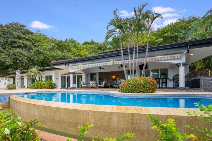 Modern house with open patio, palm trees, and a curved pool surrounded by greenery under a clear blue sky.