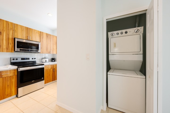 Compact kitchen with wooden cabinets, microwave, stove, and stackable washer-dryer in a closet.