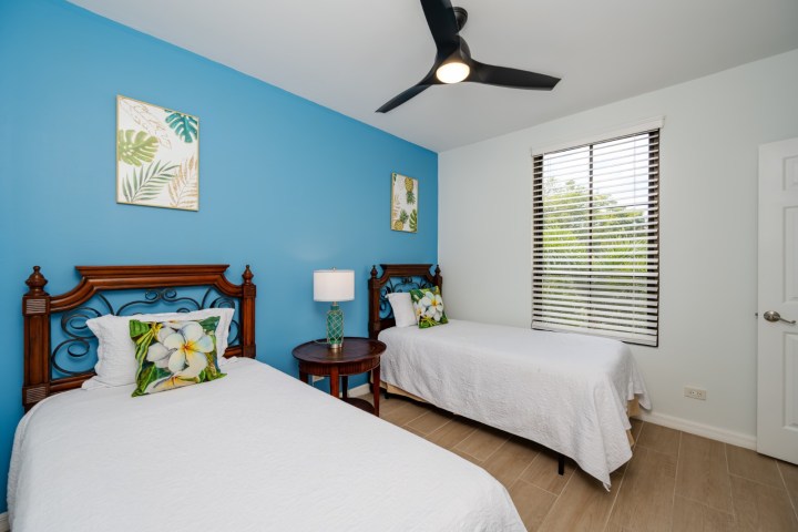Bedroom with two single beds, blue accent wall, and a ceiling fan. Tropical decor on walls and pillows.