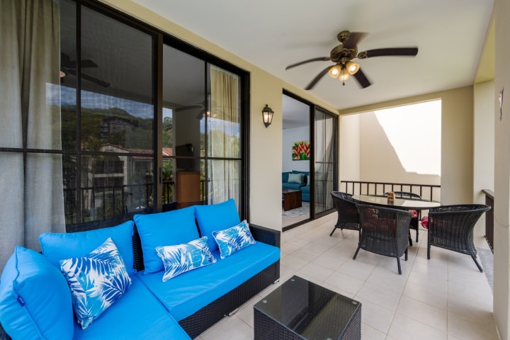 Covered balcony with blue cushioned seating, table, and ceiling fan.