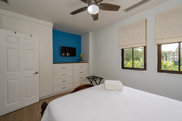Bedroom with blue accent wall, TV, dresser, ceiling fan, and two windows with outdoor view.