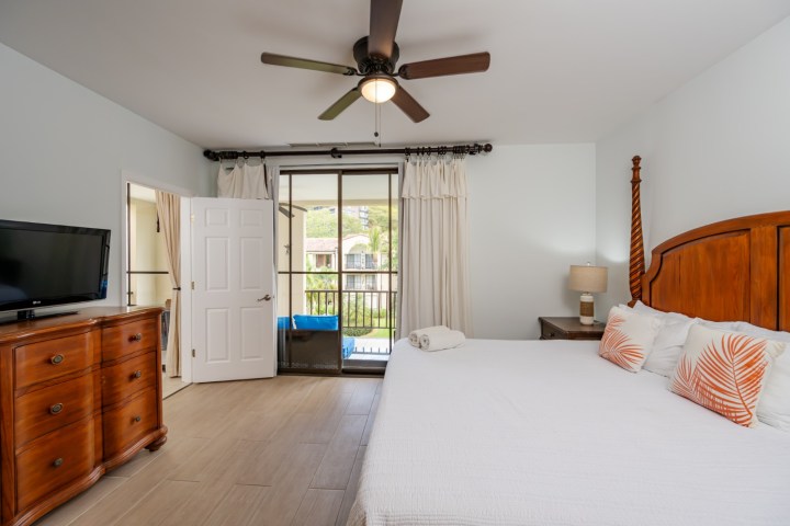 Bedroom with ceiling fan, wooden furniture, bed with pillows, TV, and open door to balcony.