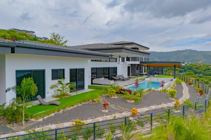 Modern house with pool, surrounded by greenery and mountains under a cloudy sky.