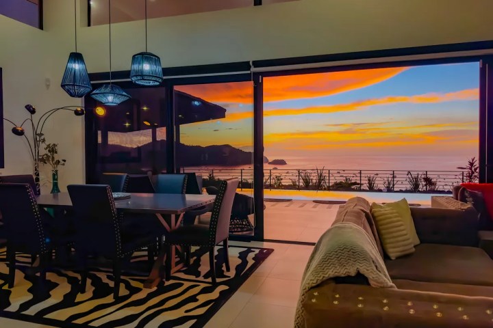 Modern dining room with ocean view, sunset through glass doors, blue pendant lights.
