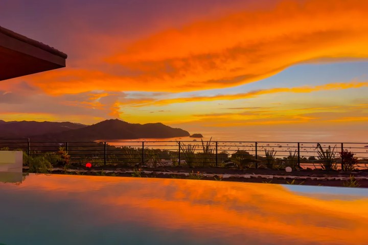 Vibrant orange sunset over ocean with silhouetted mountains and a reflective pool in the foreground.