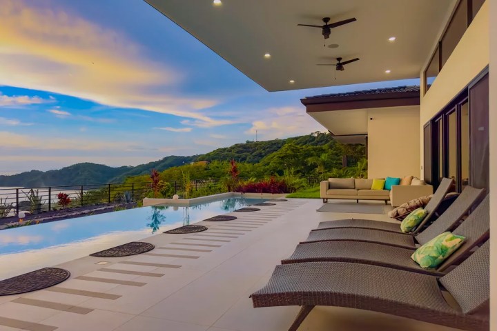 Outdoor patio with pool, lounge chairs, and mountain view under a cloudy sky at sunset.
