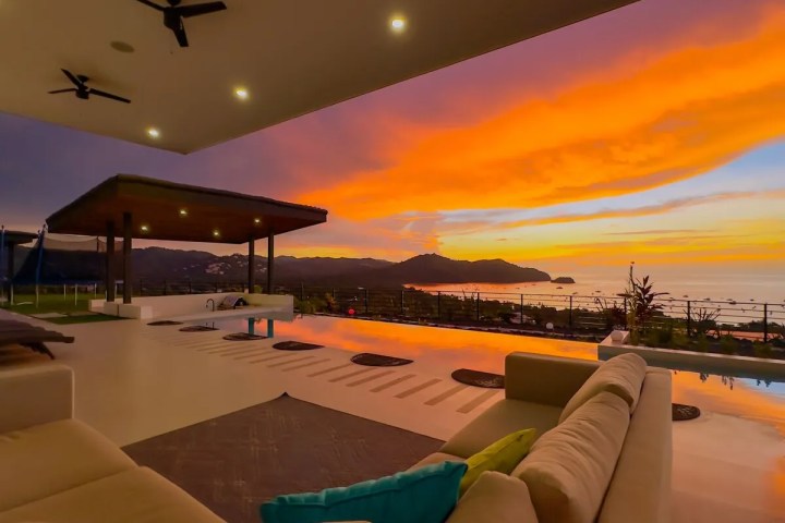 Outdoor patio overlooking ocean sunset with sofa, ceiling fans, and pool.
