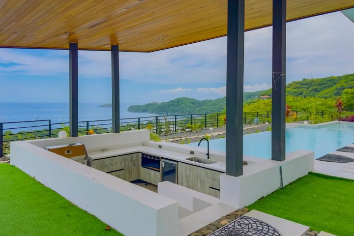 Outdoor kitchen with island, pool, and ocean view under wooden ceiling.