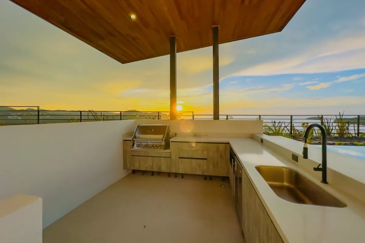 Outdoor kitchen with grill and sink under a wooden ceiling at sunset.