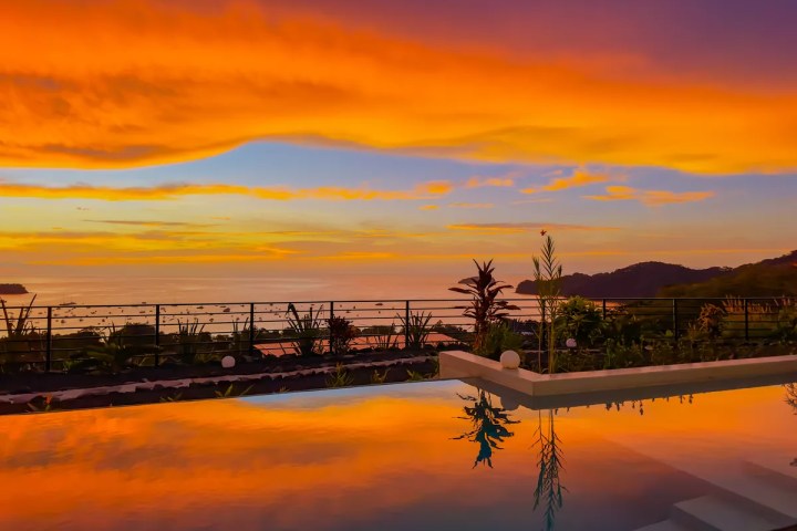 Vibrant sunset over ocean with reflections in a swimming pool and silhouette of foliage.