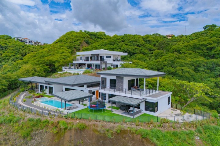 Modern hillside villa with pool and two-tiered terrace, surrounded by lush green trees under cloudy sky.