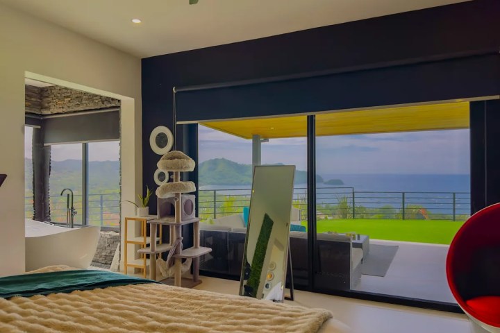 Bedroom with large window view of ocean, freestanding bathtub, cat tree, and modern decor.