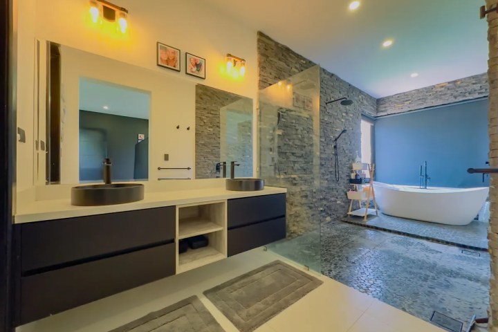 Modern bathroom with double sinks, stone shower wall, and freestanding bathtub.