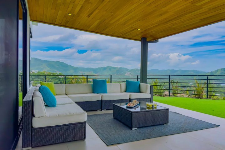 Outdoor patio with L-shaped sofa, cushions, glass-top table, and mountain view under wooden roof.