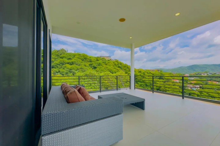Spacious balcony with wicker furniture overlooking lush green hills under a cloudy sky.