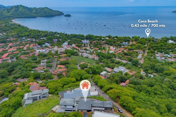 Aerial view of a town near the coast with a distance marker to Coco Beach, surrounded by green hills.