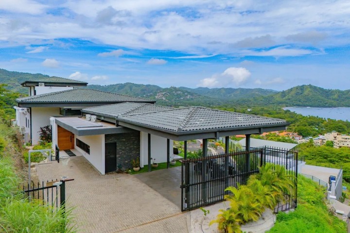 Modern hillside house with gray roofs overlooking a bay and green hills under a blue sky.