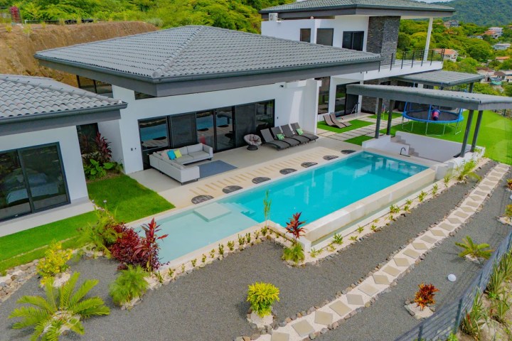 Modern white house with a pool, patio furniture, and trampoline, surrounded by greenery.