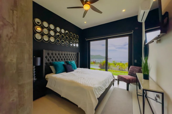 Modern bedroom with wall art, ceiling fan, large bed, and ocean view through glass doors.