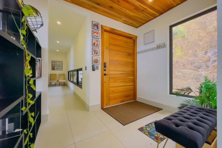Modern entryway with wooden door, tiled floor, bench, plant, and shelving with decorative items.